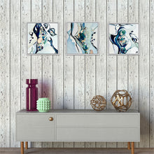 Load image into Gallery viewer, Three framed abstract paintings in blue, white, and black tones are displayed on a light gray wood-paneled wall above a gray console table with decorative vases and spheres.
