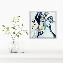 Load image into Gallery viewer, A framed abstract painting with blue, black, white, and soft peach tones hangs on a white wall above a white shelf, next to a clear glass vase holding green stems.
