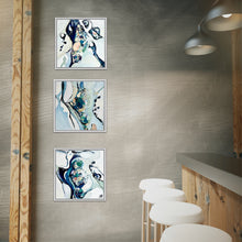 Load image into Gallery viewer, Three framed abstract wall art prints with blue, teal, and white fluid designs, displayed vertically on a textured gray wall in a modern bar or cafe setting with wooden posts, white stools, and pendant lights.
