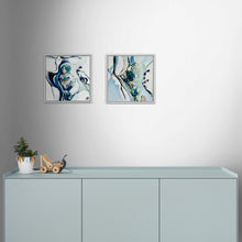 Load image into Gallery viewer, Two square abstract wall art prints in silver frames hanging side by side on a light gray wall above a pale blue cabinet, with a small potted plant and wooden toy on the cabinet surface.
